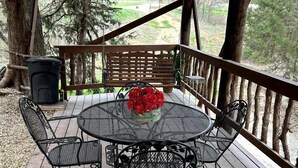 A covered outdoor deck with a round table and chairs, a ceiling fan, and a view of trees and a grassy area.