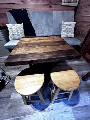 Een rustieke houten tafel met twee krukken, een grijze bank met gestreepte kussens en een houten vloer.