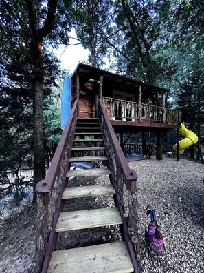 Un escalier en bois mène à une cabane dans les arbres, à laquelle on accède par un toboggan jaune.
