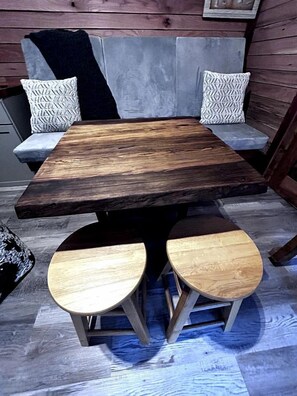 Une table en bois rustique, accompagnée de deux tabourets, un canapé gris orné de coussins à motifs et un plancher en bois.