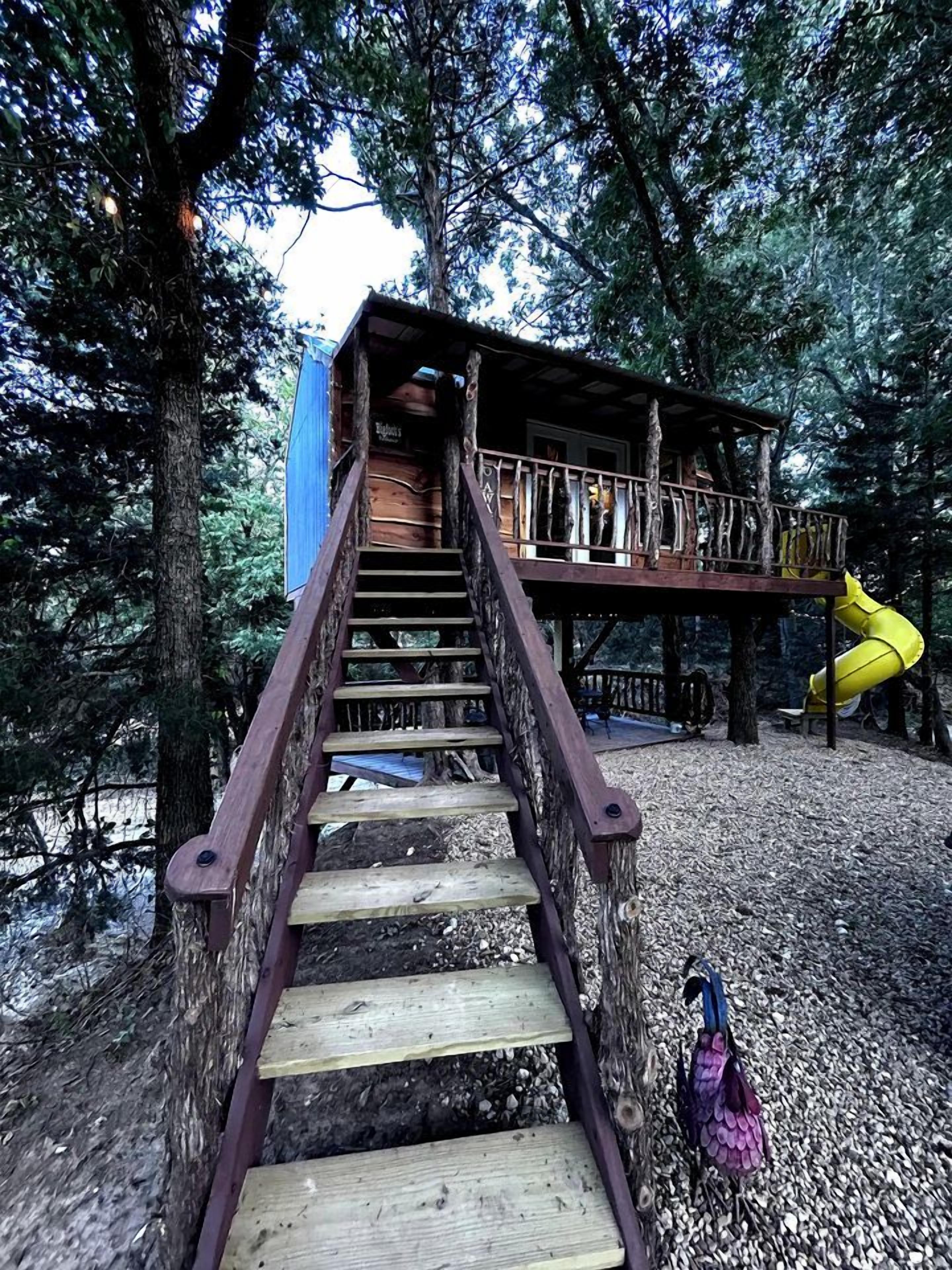 Un escalier en bois mène à une cabane dans les arbres, à laquelle on accède par un toboggan jaune.