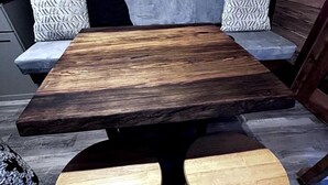A rustic wooden table with two stools, a grey sofa with patterned cushions, and a wooden floor.