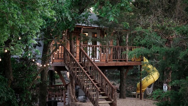 A treehouse with wooden stairs and a yellow slide, surrounded by dense foliage.