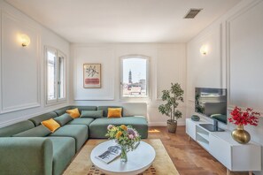A modern living room with a sectional sofa, a round coffee table with flowers, a flat-screen TV on a white stand, and a city view through large windows. - Corte delle Candele Rooftop Views (Venice)