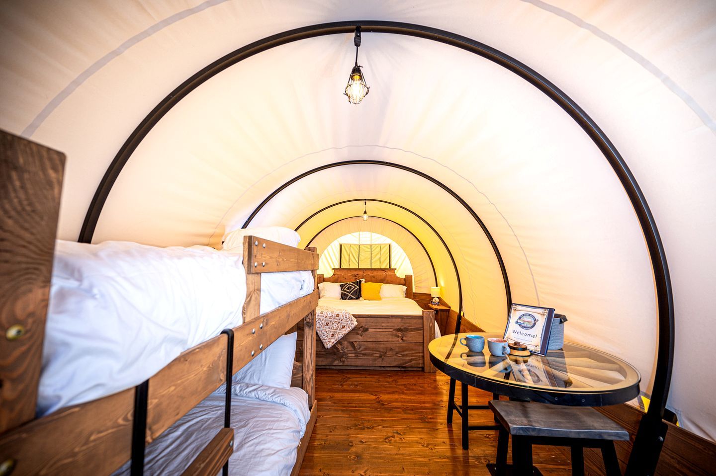 A cozy tent with two bunk beds, a small table with a cup and a book, and a welcome sign.