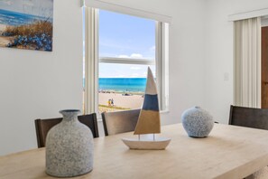 Un coin repas avec vue sur la plage, visible à travers une fenêtre, agrémenté d’une maquette de voilier et de deux vases placés sur la table.