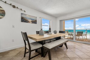 Un coin repas avec une table en bois, des chaises et un banc. Un tableau orne le mur, une fenêtre offre une vue sur l’océan et un luminaire est fixé au plafond.