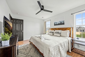 A bedroom with a bed, a ceiling fan, a desk with a plant, a window with a view, and a door. - Comfortable Retreat with Hot-Tub in Sevierville, Tennessee (Sevierville)