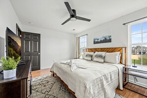 A bedroom with a bed, a ceiling fan, a desk with a plant, a window with a view, and a door. - Comfortable Retreat with Hot-Tub in Sevierville, Tennessee (Sevierville)