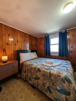 A wooden-paneled room with a bed, a nightstand, and a window with curtains. - Beachfront Cottages with Private Access & Ocean Views in North Truro, Massachusetts (North Truro)