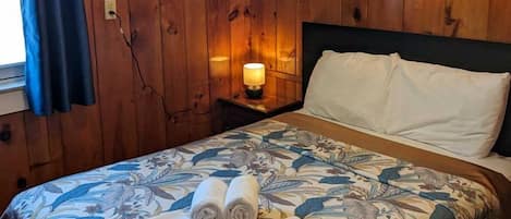 Et værelse med træpaneler, en seng med blomstret sengetøj, to sammenrullede håndklæder, et natbord med en lampe og et vindue med blå gardiner.
