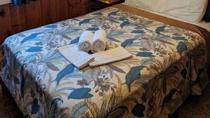 Ein Zimmer mit Holzwandverkleidung, einem Bett mit floralem Bettzeug, zwei gerollten Handtüchern, einem Nachttisch mit einer Lampe und einem Fenster mit blauen Vorhängen.