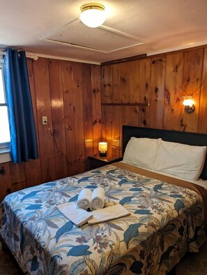 A room with wooden panel walls, a bed with floral bedding, two rolled towels, a nightstand with a lamp, and a window with blue curtains. - Beachfront Cottages with Private Access & Ocean Views in North Truro, Massachusetts (North Truro)