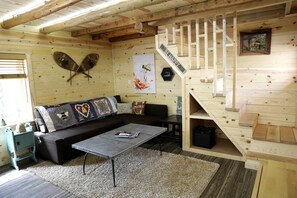 Ein gemütliches Blockhaus-Interieur mit einer Holztreppe, einem Sofa mit Steppdecke, einem Couchtisch und einem Wandventilator.