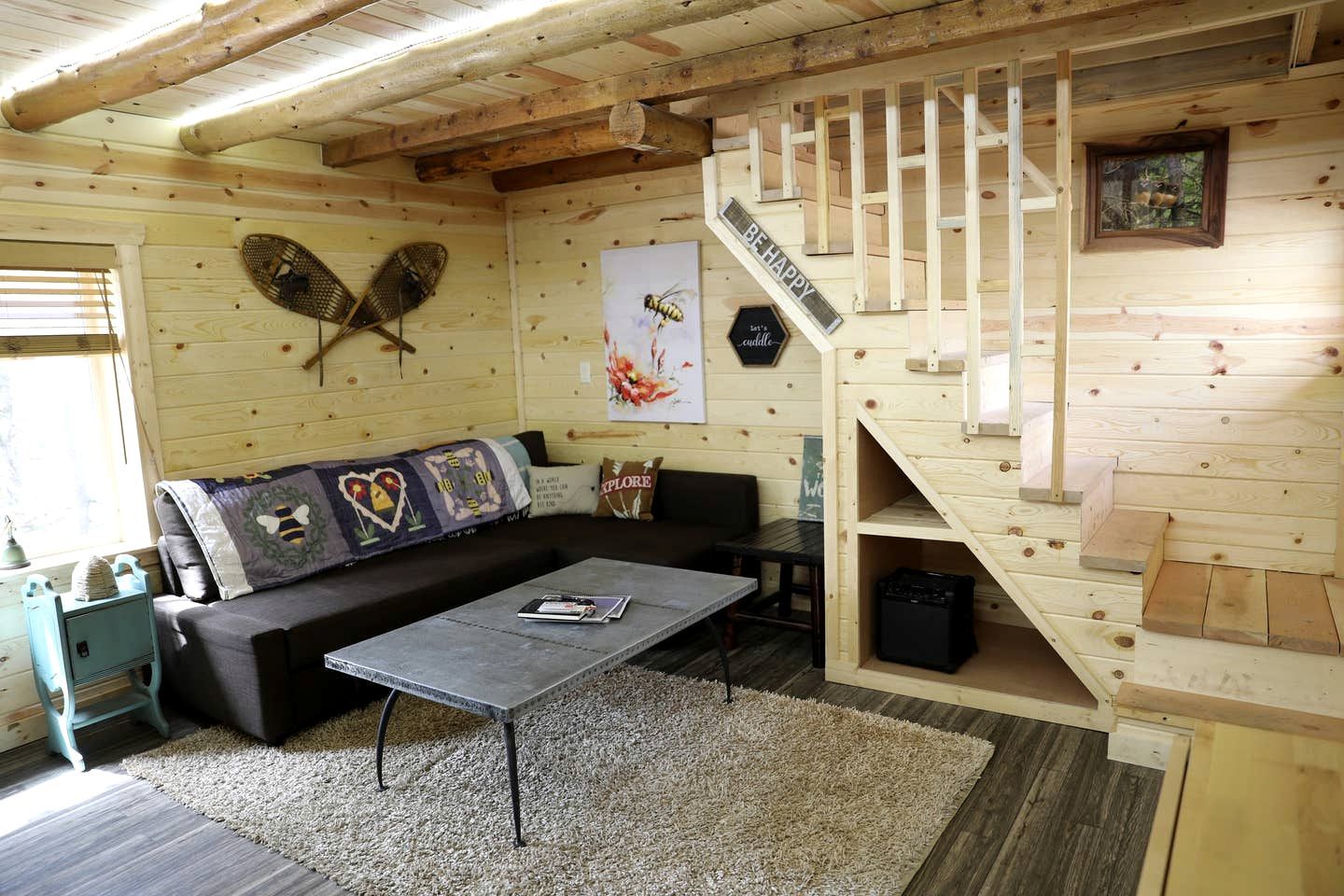 En mysig stuga med en trätrappa, en soffa med ett lapptäcke, ett soffbord och en fläkt på väggen.
