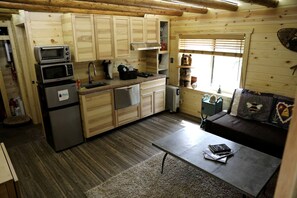 A compact kitchen with wooden cabinets, a microwave, a refrigerator, and a sink. There's a small table with a book and a chair. - Peaceful Log Cabin Perfect for a Romantic Getaway near Yellowstone, Idaho (Island Park)