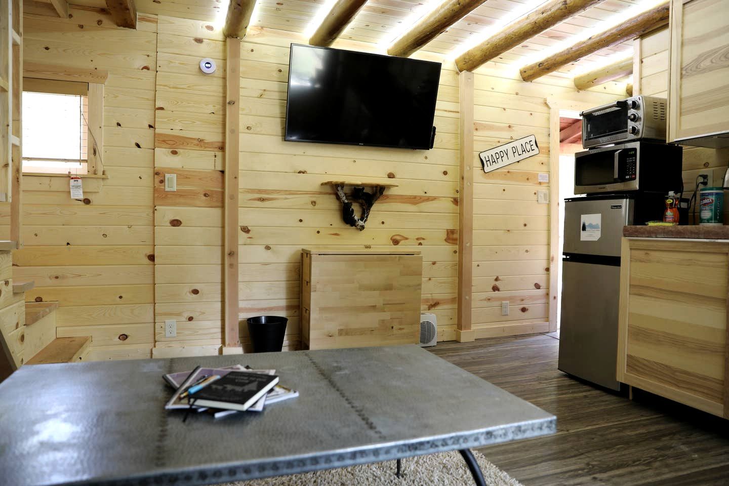 Un intérieur de chalet chaleureux avec un téléviseur, un panneau indiquant « ENDROIT IDÉAL », une cheminée, un coin cuisine et une table avec des livres.