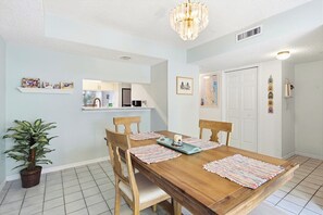 A dining area with a wooden table, chairs, a chandelier, a potted plant, and a kitchen visible through a window. - Formal dining room, warm and inviting bedrooms, and a large screened porch! (St. Augustine Beach)