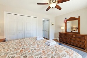 Ein Schlafzimmer mit Bett, Schminktisch, Spiegel, Deckenventilator und einer Tür zu einem anderen Raum.