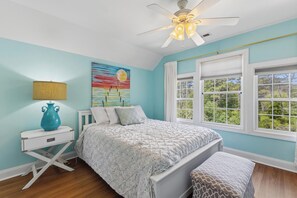 A bedroom with a bed, a nightstand, a lamp, a ceiling fan, and a window with a view of trees. - Joan and Eddie's Beach Retreat: Pet Friendly | Walking Distance to Beach (Carolina Beach)
