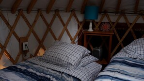Une tente dotée d’une structure en treillis de bois, un lit recouvert d’une couvre-lit à motifs bleus et blancs, une petite table de chevet avec une lampe, et un objet décoratif posé sur la table.