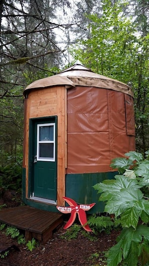 A small, wooden, dome-shaped structure with a green door and a small window, surrounded by dense forest.