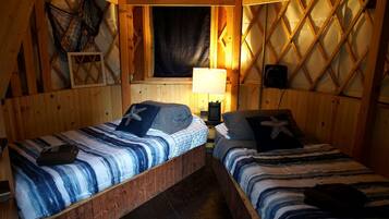 Een blokhut met twee houten stapelbedden, een raam met gordijn, een ventilator aan de muur en een klein plankje met een net.