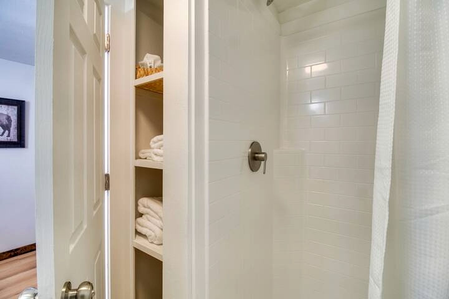 A walk-in shower with white tiled walls and a door leading to a storage area with shelves.