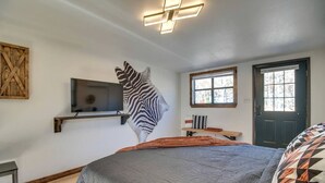 A bedroom with a bed, a mounted TV, a zebra wall art, a window, and a door. - Serene Woodland Studio Retreat with Outdoor Amenities, Near Fishing, Hiking & Gentry, AR (Gentry)