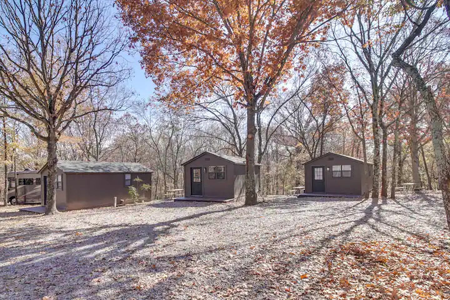 Un terrain de camping comprenant trois cabanes, entouré d’arbres et recouvert de feuilles mortes.