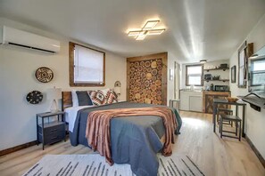 A modern bedroom with a bed, bedside tables, a wall-mounted air conditioner, a kitchen area with a microwave, and a dining table. - Serene Woodland Studio Retreat with Outdoor Amenities, Near Fishing, Hiking & Gentry, AR (Gentry)