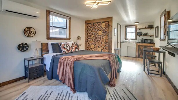 Un dormitorio con cama, mesitas de noche, aire acondicionado instalado en la pared, una ventana con persianas, un detalle decorativo en la pared, y una zona de cocina con microondas y lavamanos.