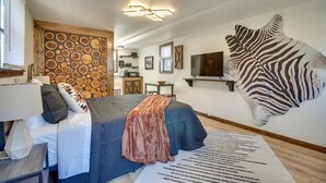 A bedroom with a bed, a large wall-mounted zebra print, a wooden wall with circular patterns, a television, and a desk.