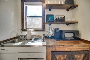Eine Küche mit Herd, Spüle, Mikrowelle und Kaffeemaschine. Über der Spüle befindet sich ein Fenster und ein Regal mit Geschirr und Besteck.