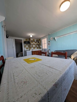 A dining area with a table covered by a lace tablecloth, wooden chairs, a kitchen in the background, and a ceiling light. - Steps to the Sand with Coastal Charm in North Truro, Massachusetts (North Truro)
