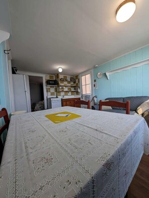 A dining area with a table covered by a lace tablecloth, wooden chairs, a kitchen in the background, and a ceiling light. - Steps to the Sand with Coastal Charm in North Truro, Massachusetts (North Truro)
