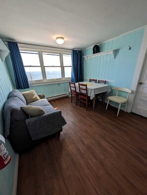 A compact room with a sofa, a dining table with chairs, a window with curtains, and a light fixture. - Steps to the Sand with Coastal Charm in North Truro, Massachusetts (North Truro)