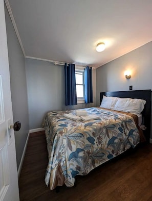 A bedroom with a bed, a window with curtains, a wall-mounted light, and a door. - Steps to the Sand with Coastal Charm in North Truro, Massachusetts (North Truro)