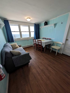 A compact room with a sofa, a dining table with chairs, a window with curtains, and a light fixture. - Steps to the Sand with Coastal Charm in North Truro, Massachusetts (North Truro)