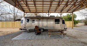 Una autocaravana estacionada sota una zona coberta, amb una taula de picnic i una cadira de fusta.