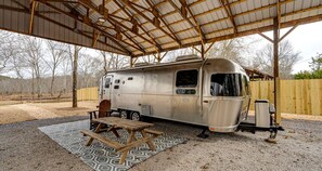 Un camping-car est stationné à l’abri, sous une zone couverte, à proximité d’une table de pique-nique et d’une chaise.