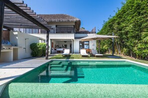 Ein modernes Haus mit Schwimmbad, überdachter Terrasse und einem strohgedeckten Gebäude.