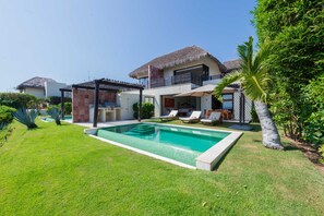 Ein modernes Haus mit Swimmingpool, einer Terrasse mit Liegestühlen und einer Struktur mit Strohdach.