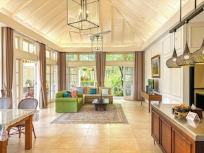 A spacious living area with a green sofa, a coffee table, and a dining table with chairs. There is a large window with curtains, a ceiling fan, and a chandelier.