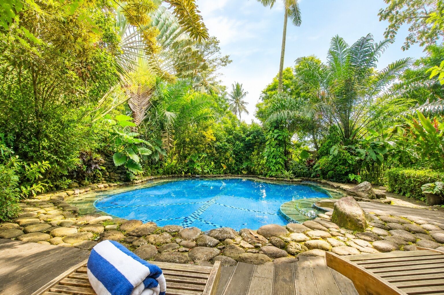 A swimming pool surrounded by tropical vegetation, with a wooden deck and a towel.