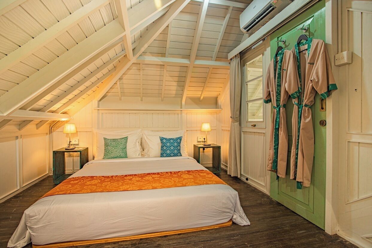 Ein Schlafzimmer mit schrägem Dach, einem Bett mit weißer Bettwäsche und einer orangefarbenen Decke, zwei Nachttischen mit Lampen sowie eine grüne Tür mit darauf liegenden Bademänteln.