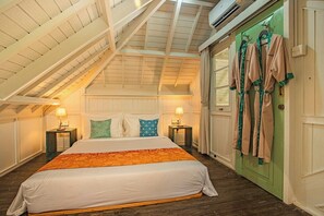Un dormitorio con techo inclinado, una cama con ropa blanca y una manta naranja, dos mesitas de noche con lámparas, y una puerta verde con batas colgadas.