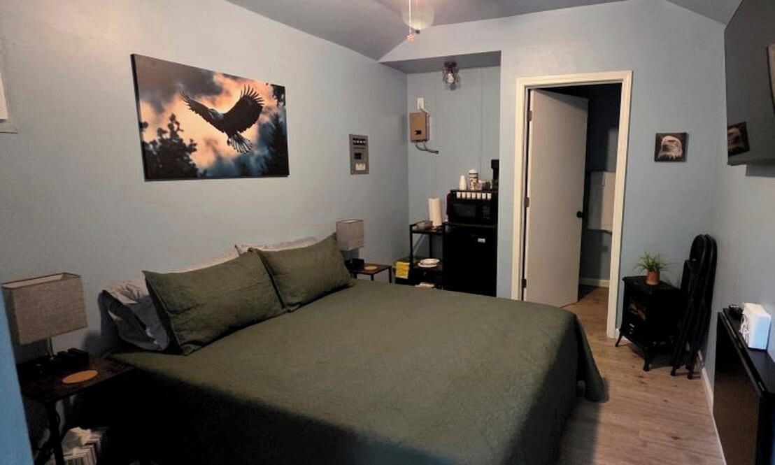 A bedroom with a bed, a nightstand, a lamp, a painting, a dresser, and a door leading to another room.