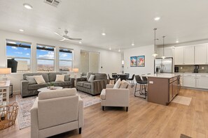 A spacious living area with sofas, a coffee table, and a dining table. There is a kitchen with white cabinets and a refrigerator. Large windows provide a view of the outdoors. - Butter by the Beach: Desert Color Lagoon + Beaches (St. George)