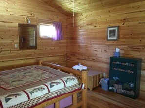 A wooden cabin room with a bed, a small table, a cabinet with a picture, and a window with curtains.